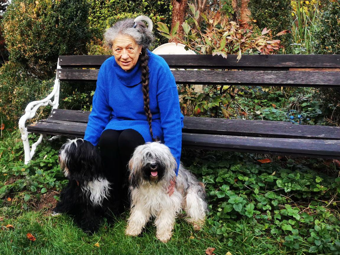 Ingrid Langer sitzt auf einer alten Gartenbank zwischen Hecken; davor stehen zwei Tibet Terrier im Gras, einer schwarz-weiß, einer hellgrau.